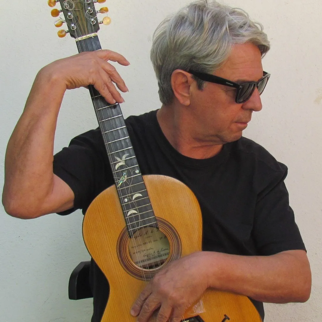 Músico de cabelo grisalho e óculos escuros segurando uma viola de pé, sobre fundo branco