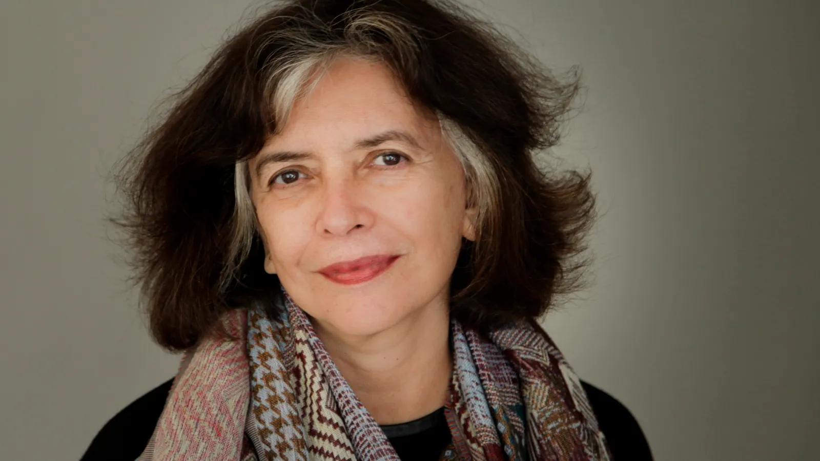 Retrato da escritora Ana Miranda, de cabelo escuro com mecha grisalha e lenço estampado, olhando para a câmera